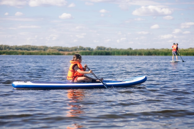 Photo little caucasian girl sitting on paddle board alone holding oar in hands and looking at other sup boarders in orange life jacket active holidays inculcation of love for sports from childhood