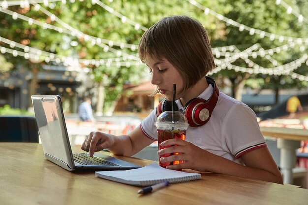 Photo little caucasian girl sitting at table at veranda of cafe studying with latop