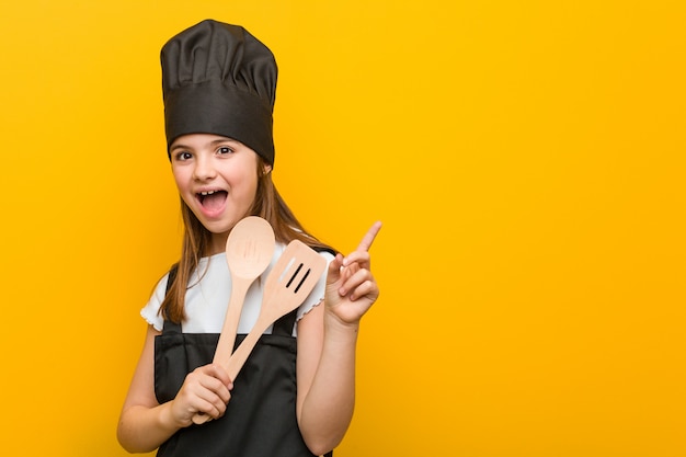 Piccola ragazza caucasica che porta un costume del cuoco unico che sorride allegramente indicando con l'indice via.