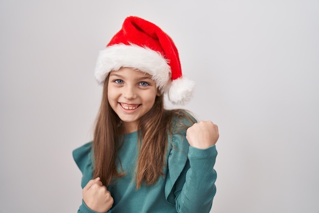 Фото Маленькая кавказская девочка в рождественской шляпе празднует удивление и изумление успеху с поднятыми руками и закрытыми глазами