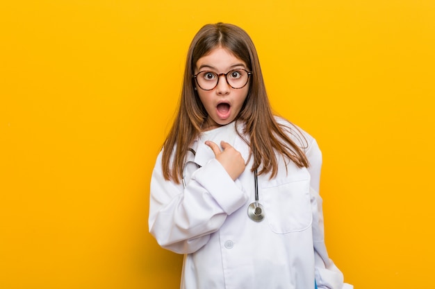 Little caucasian girl wearing a doctor costume surprised pointing at himself, smiling broadly.