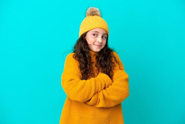 Little caucasian girl wearing winter hat isolated on blue background making doubts gesture while lifting the shoulders