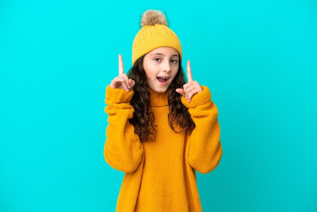 Little caucasian girl wearing winter hat isolated on blue background pointing up a great idea