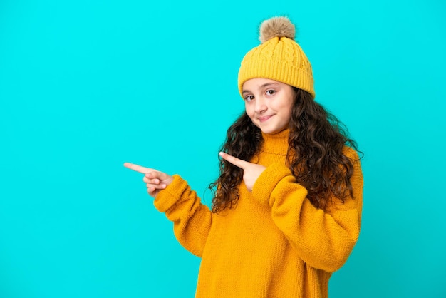Маленькая кавказская девочка в зимней шапке на синем фоне удивлена и указывает в сторону