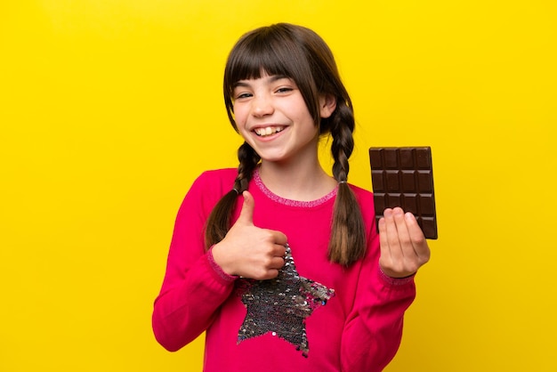 Маленькая кавказская девочка с шоколадом на желтом фоне показывает большой палец вверх