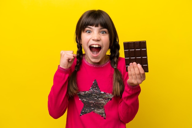 Little caucasian girl with chocolat isolated on yellow background celebrating a victory in winner position