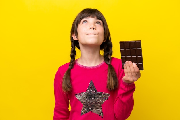 Little caucasian girl with chocolat isolated on yellow background and looking up