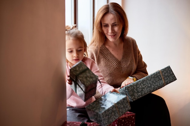 Little caucasian girl with mother unpacking gift boxes at home near window Happy family christmas concept