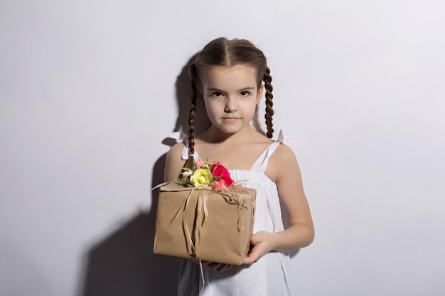 Little caucasian girl with pigtails and dark eyes standing in a dress on a white background holds a gift in hands
