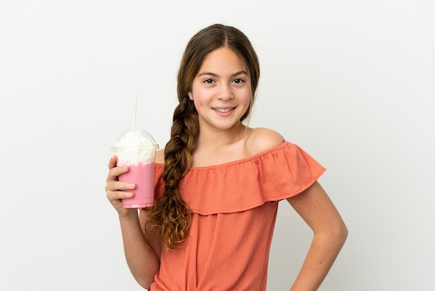 Little caucasian girl with strawberry milkshake isolated on white background posing with arms at hip and smiling