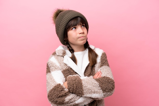 Little caucasian girl with winter jacket isolated on pink background making doubts gesture while lifting the shoulders