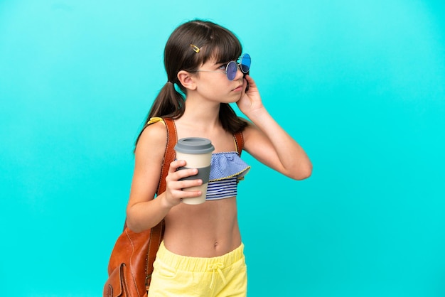 Little caucasian kid going to the beach isolated on blue background holding coffee to take away and a mobile