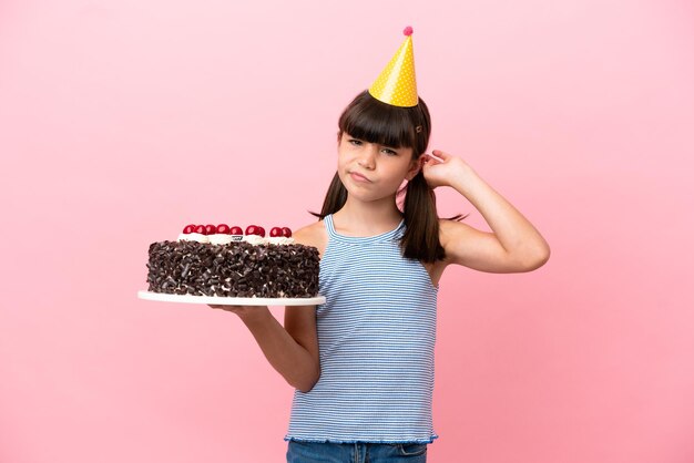 Little caucasian kid holding birthday cake isolated in pink background having doubts