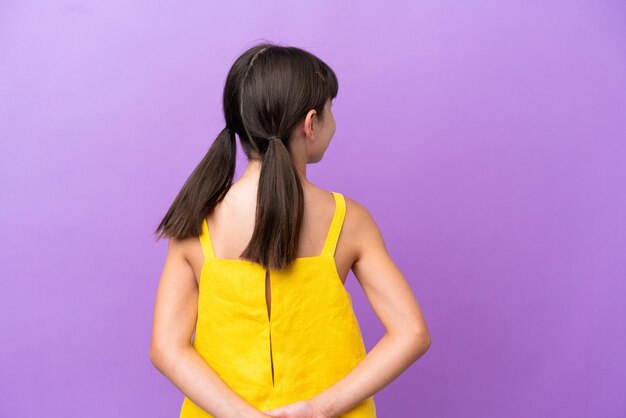 Little caucasian kid isolated on purple background in back position and looking back