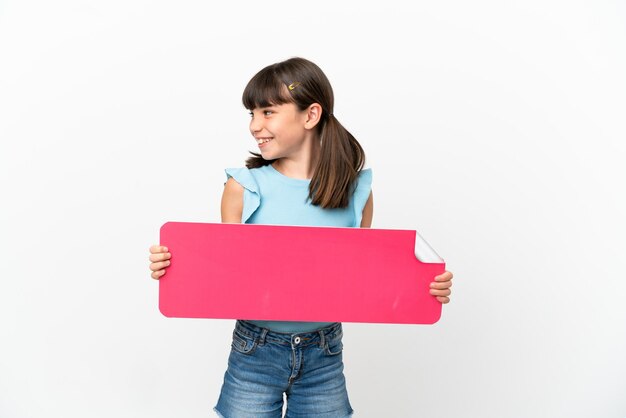 Little caucasian kid isolated on white background holding an empty placard and looking side