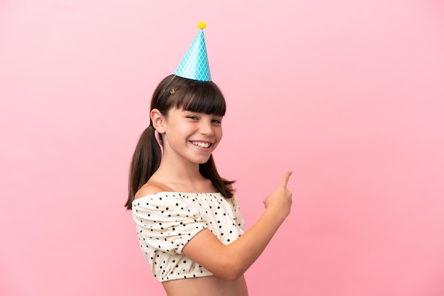 Little caucasian kid with birthday hat isolated on pink\
background pointing back