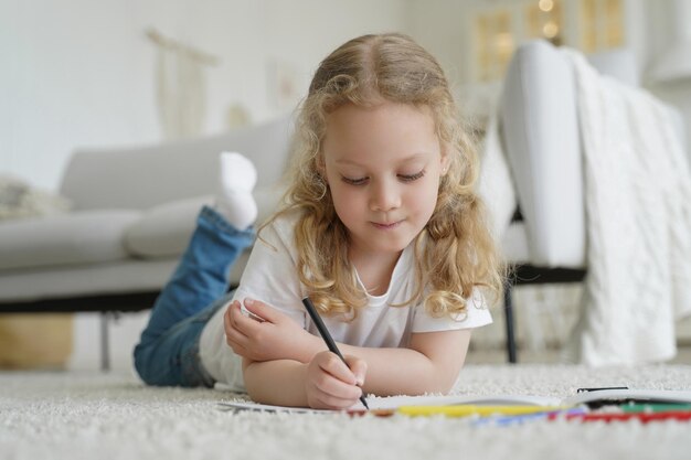 Little caucasian preschooler child girl drawing with crayon lying on warm floor Creative art hobby