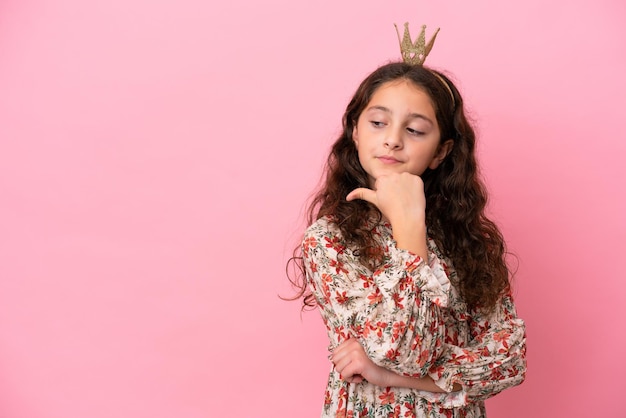 Little caucasian princess with crown isolated on pink background pointing to the side to present a product