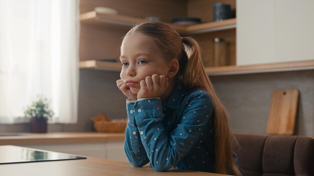 Little caucasian sad upset child girl sitting at home kitchen alone offended kid schoolgirl feeling