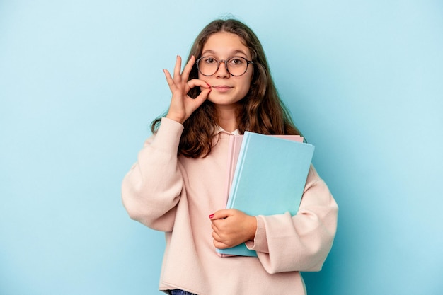 Piccola studentessa caucasica che tiene libri isolati su sfondo blu con le dita sulle labbra mantenendo un segreto.