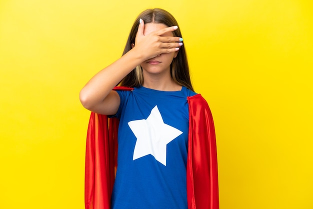 Little caucasian superhero girl isolated on yellow background\
covering eyes by hands do not want to see something