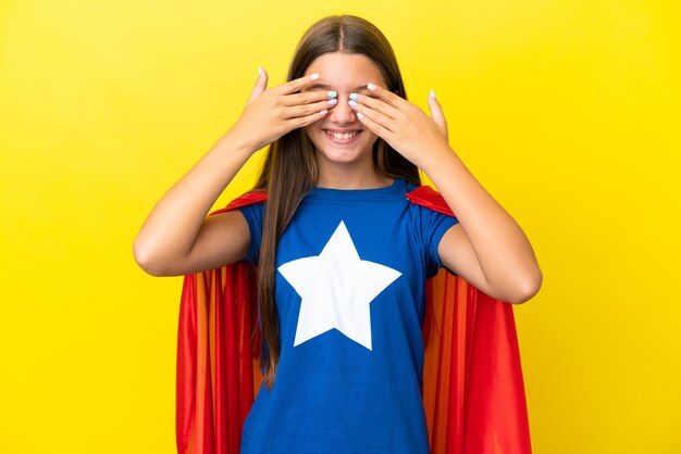 Little caucasian superhero girl isolated on yellow background covering eyes by hands