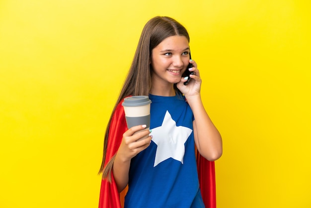 Little caucasian superhero girl isolated on yellow background holding coffee to take away and a mobile