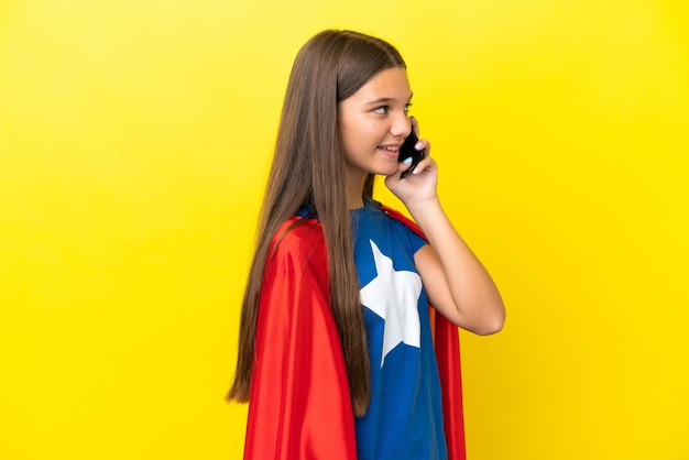 Little caucasian superhero girl isolated on yellow background keeping a conversation with the mobile phone