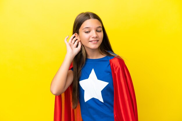 Little caucasian superhero girl isolated on yellow background listening to something by putting hand on the ear