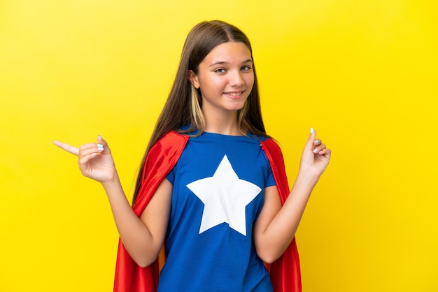 Foto piccola ragazza caucasica supereroe isolata su sfondo giallo che punta il dito verso i laterali e felice