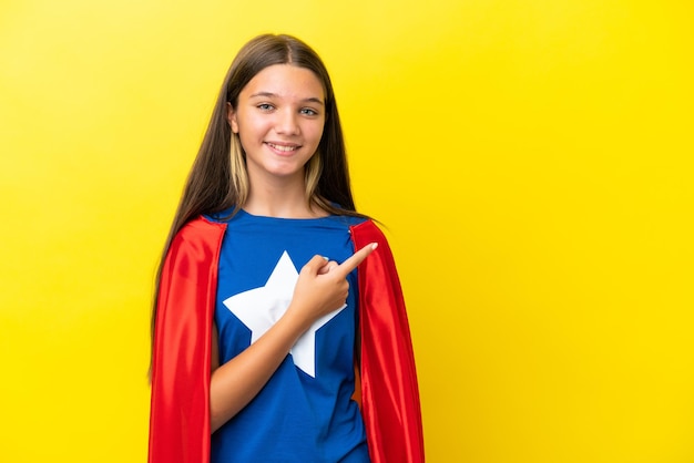 Little caucasian superhero girl isolated on yellow background pointing to the side to present a product