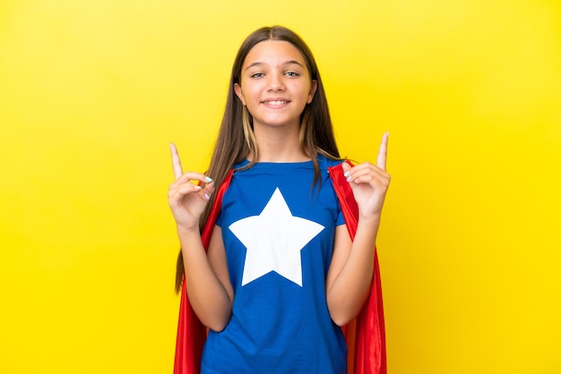 Little caucasian superhero girl isolated on yellow background pointing up a great idea