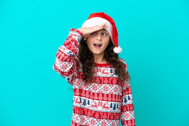 Little caucasian with christmas hat isolated on blue background with surprise and shocked facial expression