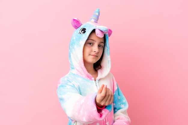 Little caucasian woman wearing a unicorn pajama isolated on pink background making money gesture
