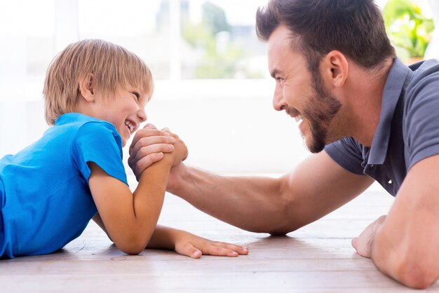 Little champion. Side view of happy father and son competing in arm wrestling while both lying on the hardwood floor