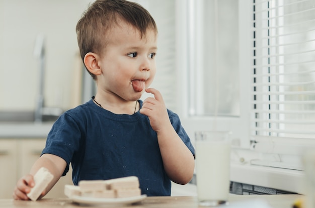 A little charming boy eats waffles in the kitchen and drinks milk