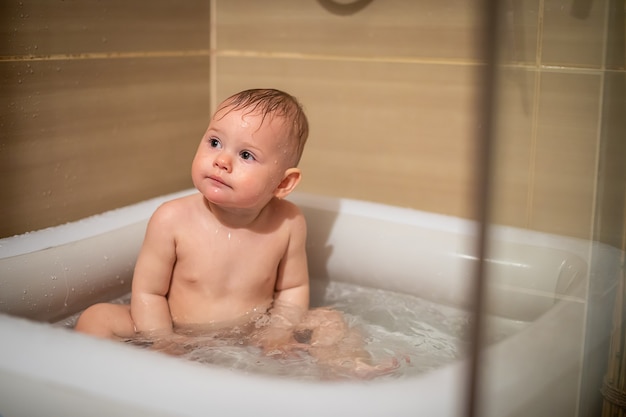 Little charming girl babe plays in the water in a small inflatable baby pool located in the shower stall in the bathroom, baby girl smiling and having fun