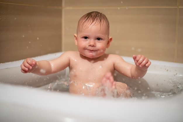 Маленькая очаровательная девочка-младенец играет в воде в небольшом надувном детском бассейне, расположенном в душевой кабине в ванной комнате, девочка улыбается и веселится