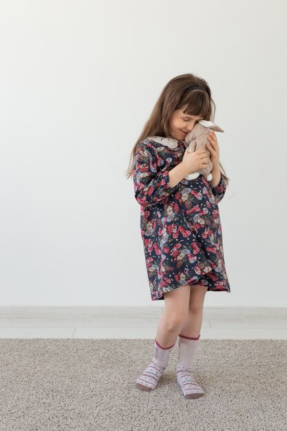 Little charming girl in a flower dress gently embraces her favorite toy bunny standing on a white wall