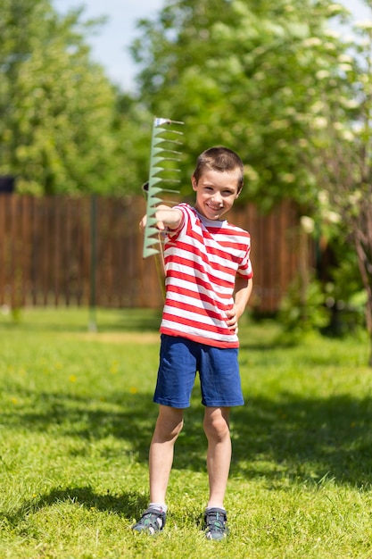 A little cheerful boy stands and holds a rake in his hand, ready to work