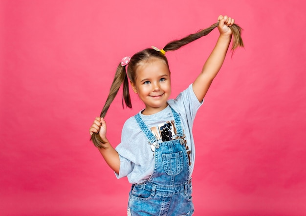 Маленькая жизнерадостная девочка в джинсовом комбинезоне тянет волосы в разные стороны. дети.