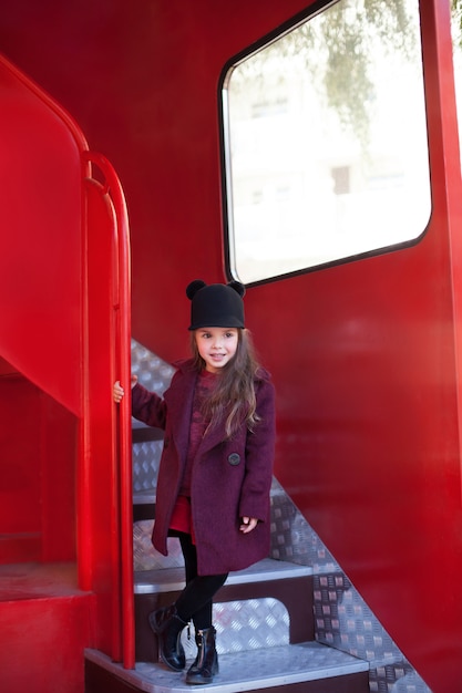 Little cheerful girl near the red English bus in a beautiful coat and a hat. Child's journey. School bus.  London red bus. Spring.  With the International Women's Day. Since March 8!