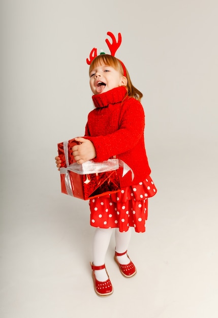 La piccola ragazza allegra con un maglione rosso tiene una scatola regalo rossa su sfondo bianco.