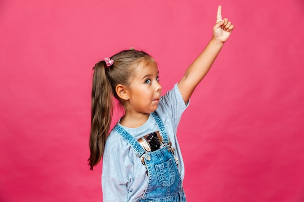 Маленькая жизнерадостная девочка показывает пальцем в сторону и вверх на розовом фоне. дети.
