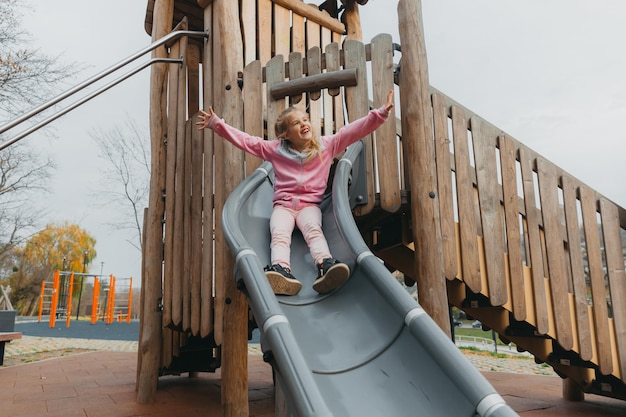 A little cheerful girl with long blond hair goes down the\
children\'s slide.