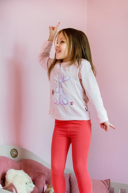 маленькая веселая девочка с длинными светлыми волосами прыгает на детской уютной кровати в своей комнате