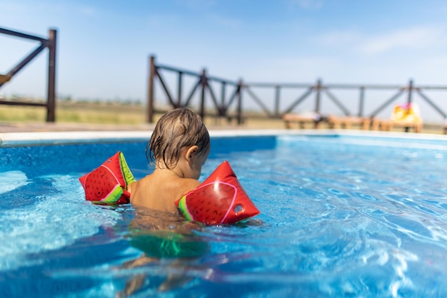 밝은 부풀릴 수 있는 완장을 한 밝고 행복한 소년 유아는 따뜻한 여름 태양 아래 맑고 투명한 물을 가진 아이들을 위해 파란색 수영장에서 수영합니다