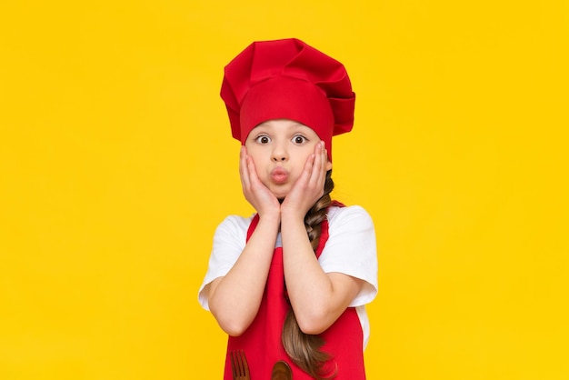 The little chef girl puffed out her cheeks Cooking for children A child in a red apron and a cap on a yellow isolated background