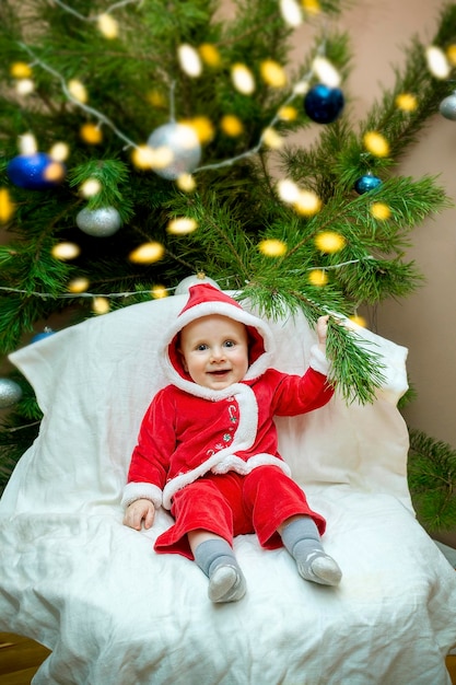 Little child baby boy sitting in chair near christmas tree in living room in red santa claus costume