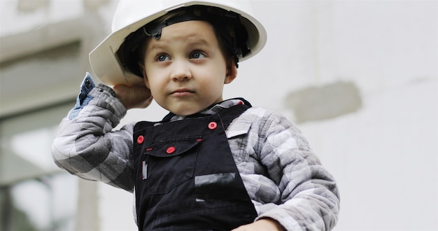 A little child builder in a white helmet sits on the scaffolding Baby boy portrait